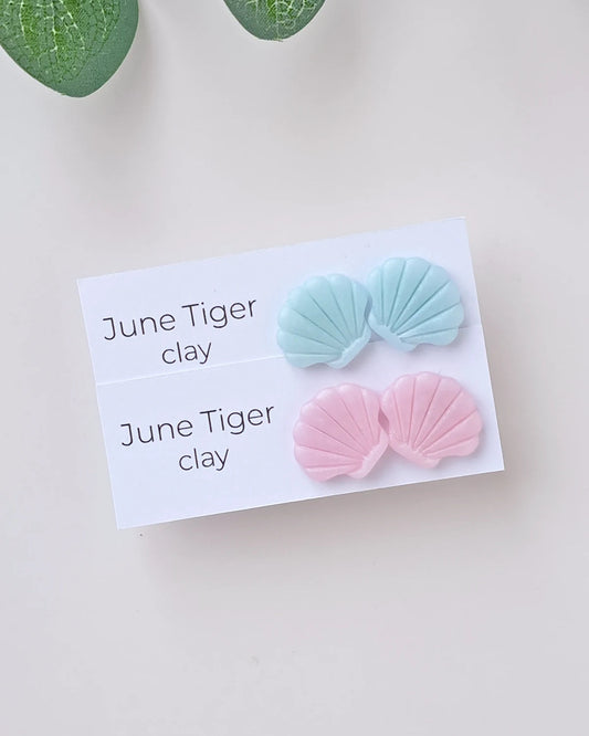 Pair of pink and blue shell-shaped earrings on a card labeled 'June Tiger clay' with green leaves in the corner.